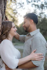 A couple stands closely by a tree, gazing at each other with soft smiles, surrounded by the warm glow of sunlight filtering through nearby branches.