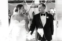 The newlyweds, smiling at each other, hold hands as they walk down the aisle lined with string lights and soft floral decorations.