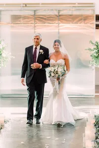 The bride, in a strapless lace wedding gown and veil, walks arm-in-arm with her father down the aisle adorned with candles and greenery, both looking forward with calm expressions.