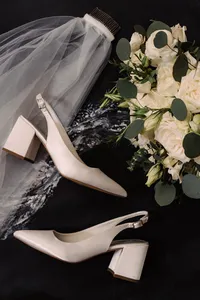 A stylish flatlay of bridal accessories, including white slingback heels, a veil, and a bouquet of white roses with eucalyptus, set against a dark background for a sophisticated touch.