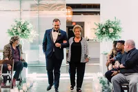 Groom walks down the aisle arm-in-arm with a smiling woman, possibly a family member, as guests watch with warm smiles.