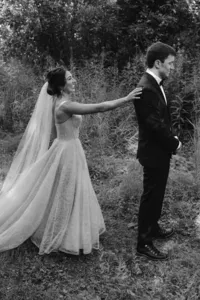 Black-and-white photo of the smiling bride reaching to tap the groom’s shoulder before their first look.