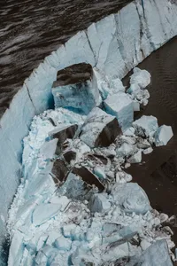 Aerial view of jagged blue-white glacier ice breaking apart along a dark glacial pool in the Alaska mountains.