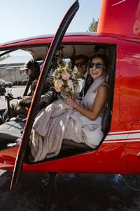 Smiling bride and groom sit inside a red helicopter, the bride holding her bouquet as they prepare for their elopement flight.