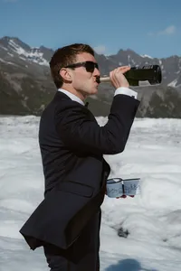 A groom in a black suit and sunglasses drinks from a champagne bottle while holding two cups on a remote Alaskan glacier, with snow-covered mountains in the background.