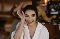 Smiling bride in pale pink pajamas touches her face after having her makeup done for the Alaska elopement.