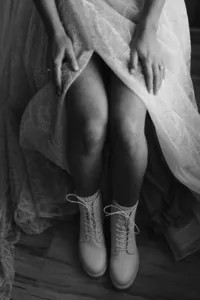 Black-and-white close-up of the seated bride lifting her lace wedding dress to reveal white lace-up boots.