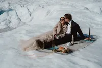 Groom kisses the smiling bride during their glacier picnic, with mugs, champagne and her bouquet arranged beside them.