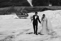 Black-and-white photo of the newlyweds holding hands as they walk across an Alaska glacier, with their helicopter behind them.