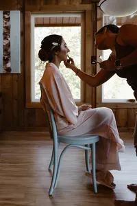 Bride in pale pink pajamas sits by cabin windows as a makeup artist applies lipstick before the Alaska elopement.