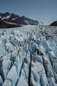 Aerial view of jagged blue-white glacier ice fractured into deep crevasses, with snow-capped Alaska mountains beyond.
