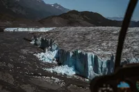 Aerial view of a towering blue ice wall along an Alaska glacier, with broken ice scattered below and rugged mountains beyond.
