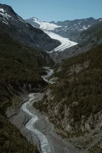 A winding glacial river cuts through a rugged Alaska valley, flowing down from a massive glacier between steep mountains.
