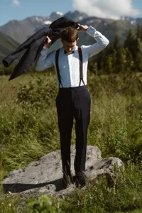 Groom in a white shirt and dark suspenders slips on his suit jacket while standing on a rock in a grassy Alaska landscape.