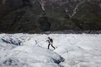 Groom runs across the uneven surface of an Alaska glacier carrying a picnic basket, with steep mountains behind him.