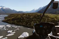View from a helicopter over a glacial river dotted with ice, surrounded by dense Alaska forest and distant mountains.