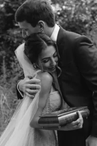 Black-and-white photo of the bride smiling as she embraces the groom, holding a leather-bound vow book.