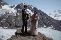 Newlyweds toast with champagne on a rocky overlook above a vast Alaska glacier, with snow-covered mountains behind them.