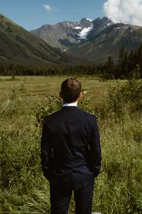 Groom in a dark suit stands in a grassy Alaska valley, looking toward forested mountains and distant glaciers.