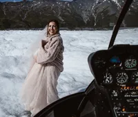 Smiling bride in a cozy coat stands on an Alaska glacier beside the helicopter, with rugged mountains rising behind her.