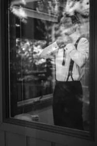 Black-and-white photo of the groom adjusting his tie while wearing a white dress shirt and dark suspenders.