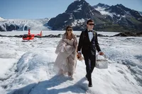 Newlyweds walk across an Alaska glacier carrying a picnic basket and bouquet, with their red helicopter and rugged mountains behind them.