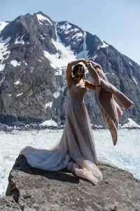 Bride in sunglasses lifts a blanket over her head while standing on a rocky outcrop above an Alaska glacier.