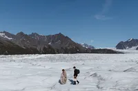 Newlyweds cross the vast white expanse of an Alaska glacier beneath rugged mountains and a clear blue sky.