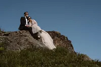 Newlyweds sit together on a rocky mountainside, the bride leaning against the groom beneath a clear blue Alaska sky.