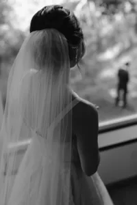 Black-and-white photo of the veiled bride looking outside, with the groom blurred in the distance awaiting their first look.