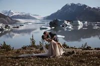 Groom dips and kisses the bride beside an iceberg-filled Alaska glacial lake, with a sprawling glacier and mountains beyond.