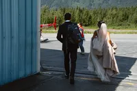 Bride and groom walk from the hangar toward a red helicopter, carrying their gear for the next part of their Alaska elopement.