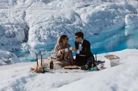 Newlyweds toast with mugs during a picnic on the Alaska glacier, surrounded by dramatic blue ice.
