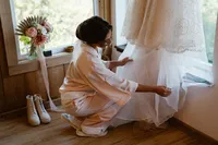 Bride in pale pink pajamas kneels beside her wedding dress, gently arranging its layered tulle skirt before getting dressed.