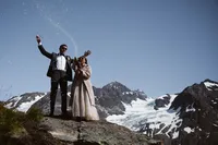 Newlyweds celebrate with a champagne spray on a rocky overlook above a sprawling Alaska glacier and snow-covered mountains.