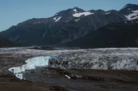 Expansive Alaska glacier stretches beneath rugged, snow-dusted mountains, with blue ice exposed along its edge.