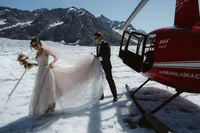 Groom holds the bride’s flowing train as they step away from a red helicopter onto an Alaska glacier surrounded by mountains.