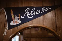 Vintage triangular pennant reading “Alaska” hangs on a wood-paneled wall above a round mirror.