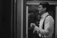 Black-and-white photo of the smiling groom getting ready in a white dress shirt and dark suspenders.