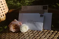 Hand-addressed envelopes and blush and white flowers rest on a woven table in warm sunlight.
