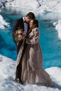 A newlywed couple embraces on a remote Alaskan glacier, laughing together beside vivid turquoise glacial water, with the bride holding a bouquet and wearing a gray fur coat over her wedding dress.