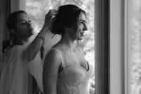 Black-and-white photo of the bride smiling as someone secures her veil into her updo beside a cabin window.