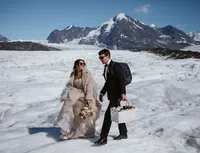 Newlyweds carry a bouquet and picnic basket across an Alaska glacier, with snow-covered mountains rising behind them.