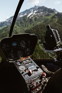 View from inside the helicopter cockpit, with flight instruments in the foreground and rugged green Alaska mountains beyond.