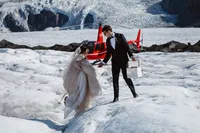 Groom helps the bride across uneven glacier ice as she gathers her dress, with their red helicopter behind them.