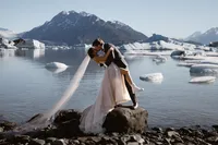 Groom dips and kisses the bride on a rocky shore, surrounded by floating icebergs and dramatic Alaska mountains.