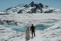 Newlyweds hold hands on a vast Alaska glacier, facing turquoise meltwater and dramatic snow-covered mountains.