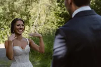 Smiling bride gestures excitedly toward the groom during their first look in a sunlit Alaska forest.