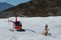 Bride and groom walk across a remote Alaska glacier beside the red helicopter that carried them into the mountains.