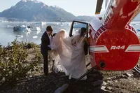 Groom helps gather the bride’s flowing dress as she climbs into a red helicopter beside an iceberg-filled Alaska lake.